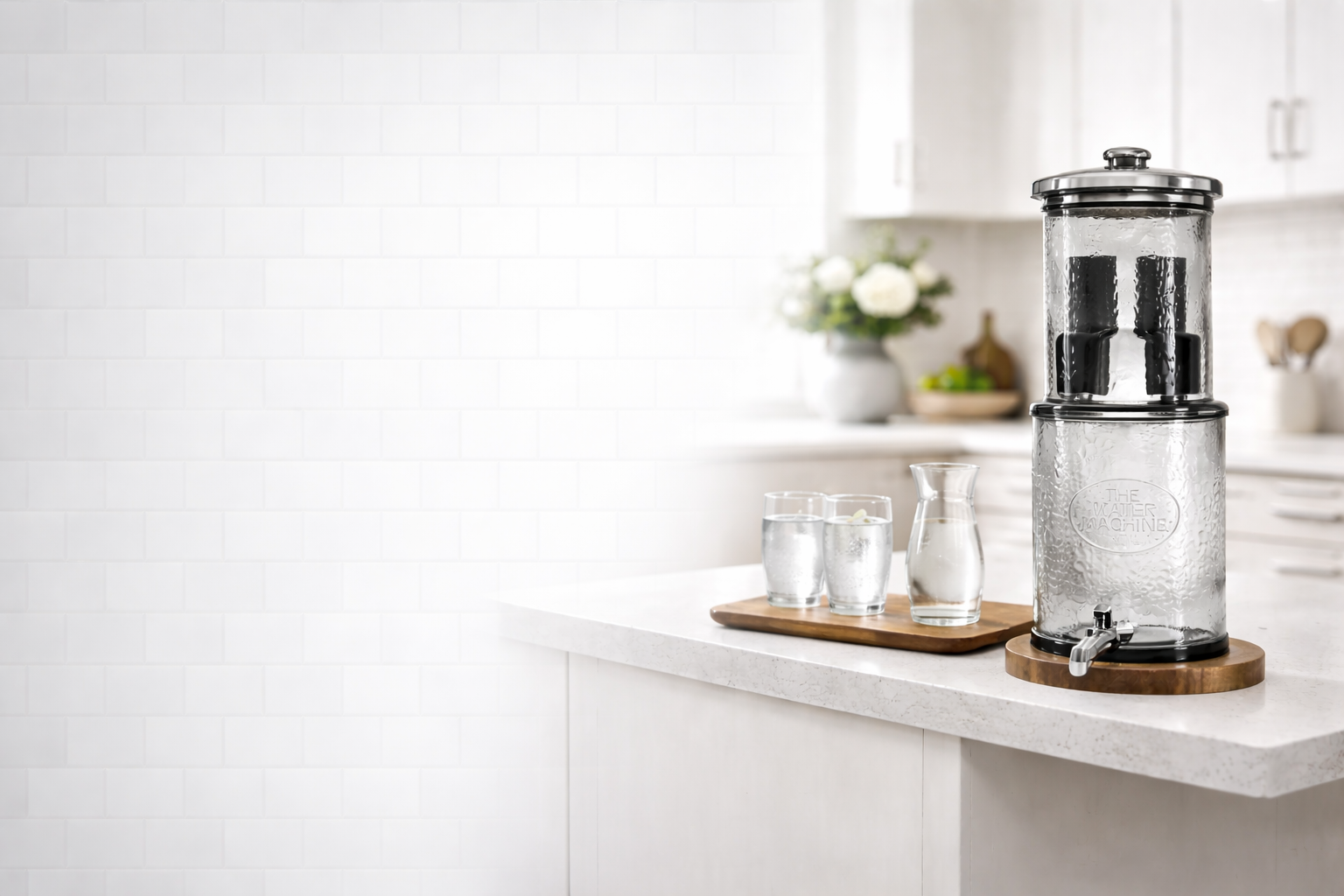 A bright white kitchen counter with the glass water purifier standing on a wooden base, two poured glasses and a carafe on a wooden board beside it.